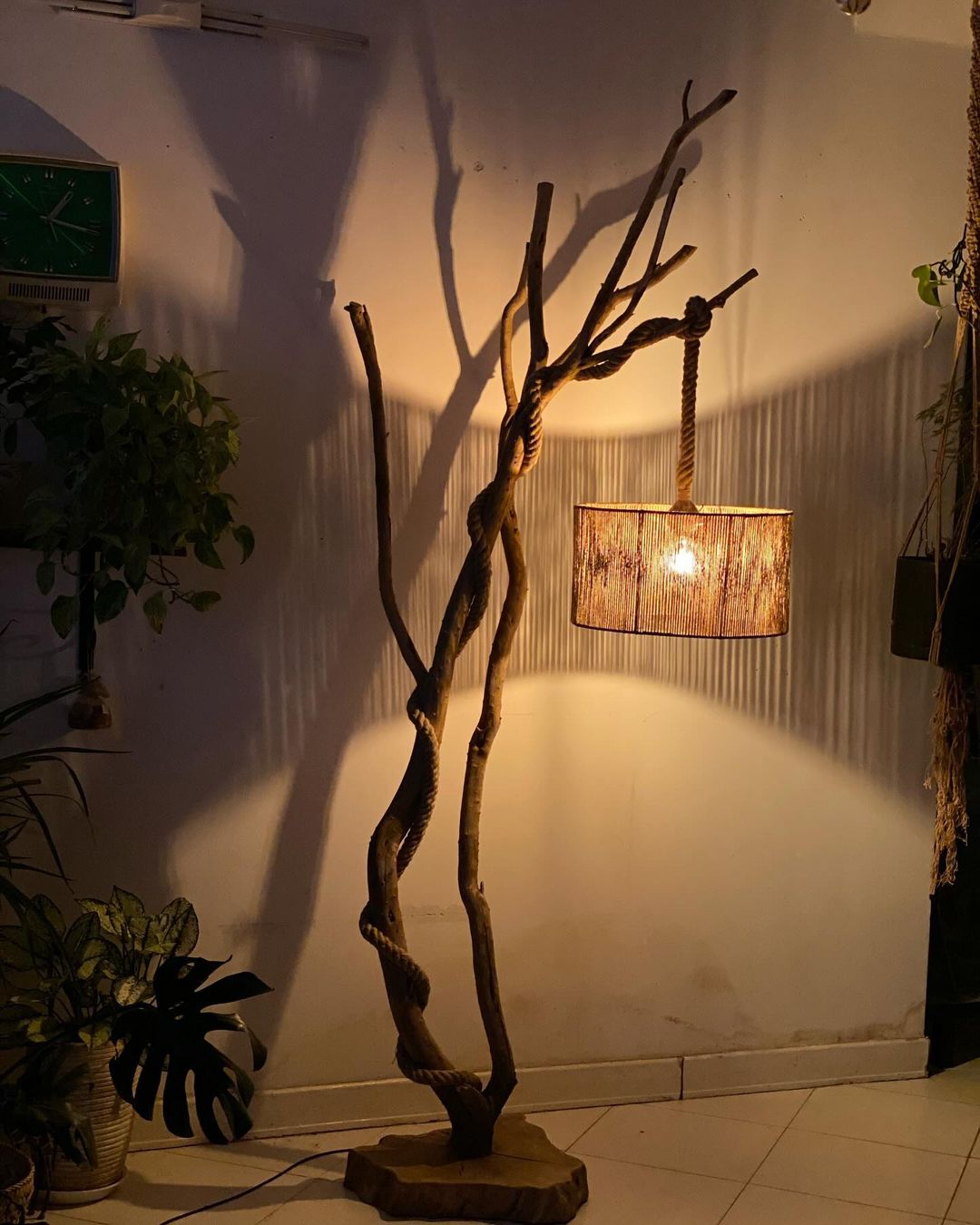 A rustic and artsy lamp stand created from twisted branches, with a rope-accented hanging lampshade, casting a soft glow in a room adorned with green plants.