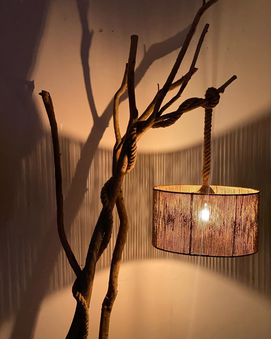 A warm and inviting lampshade suspended by intertwined branches casts playful shadows on a corrugated backdrop, exuding rustic charm.