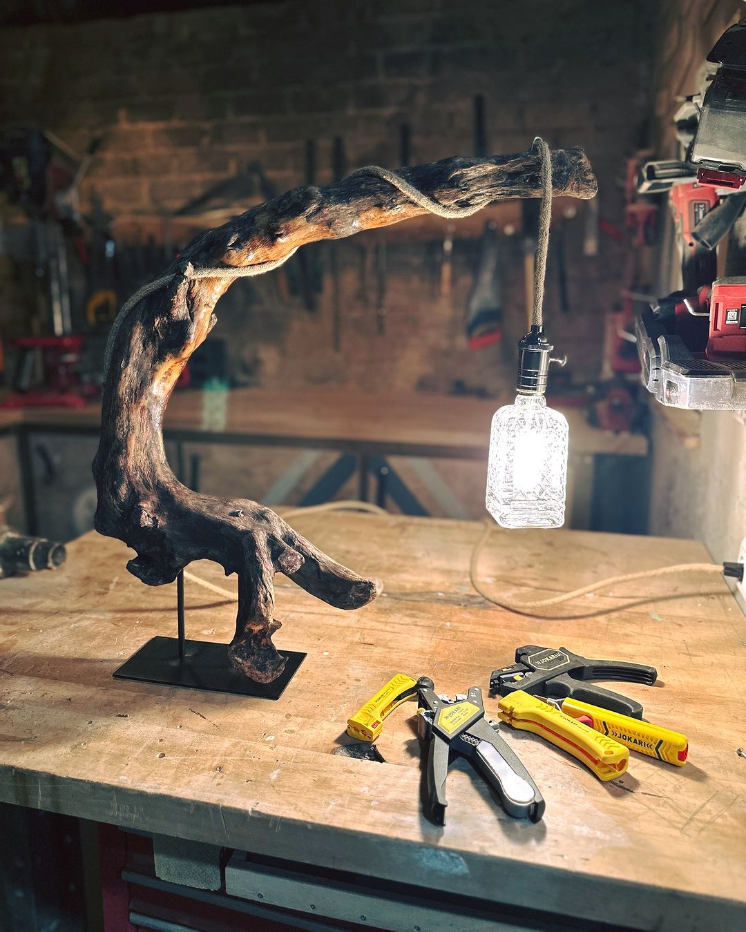 A uniquely crafted wooden lamp with an Edison bulb, rising from a sturdy black base, resting on a worn craftsman's table surrounded by tools.