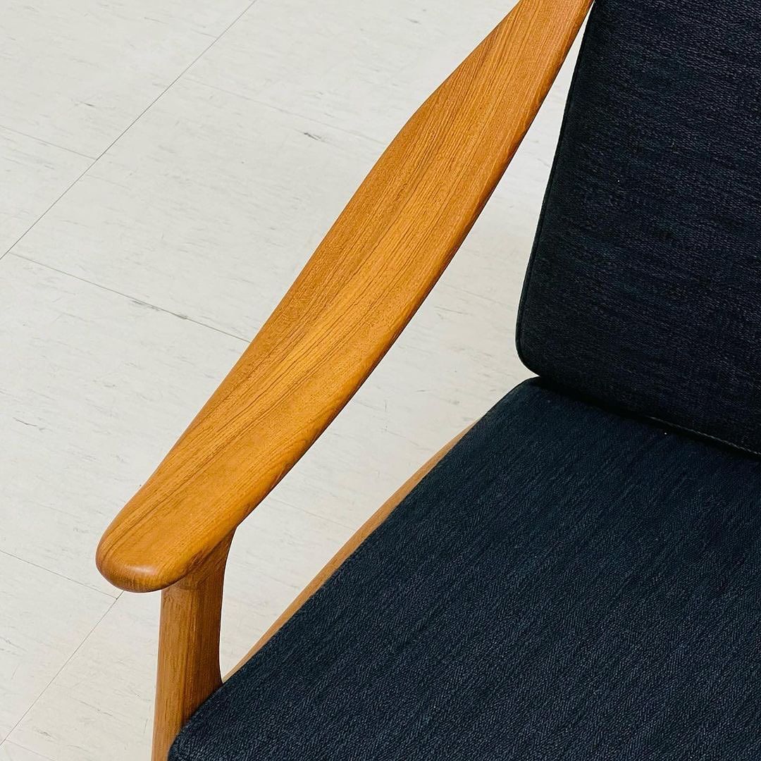 A modern minimalist chair featuring a smooth wooden armrest and a dark textured fabric seat.