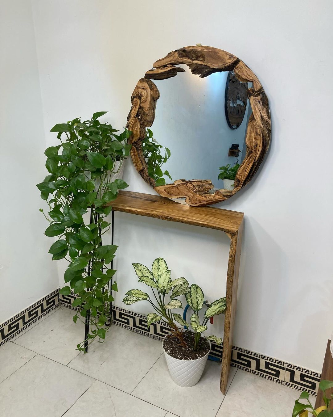 A rustic wooden-framed circular mirror creates a focal point above a sleek console table, flanked by lush greenery.