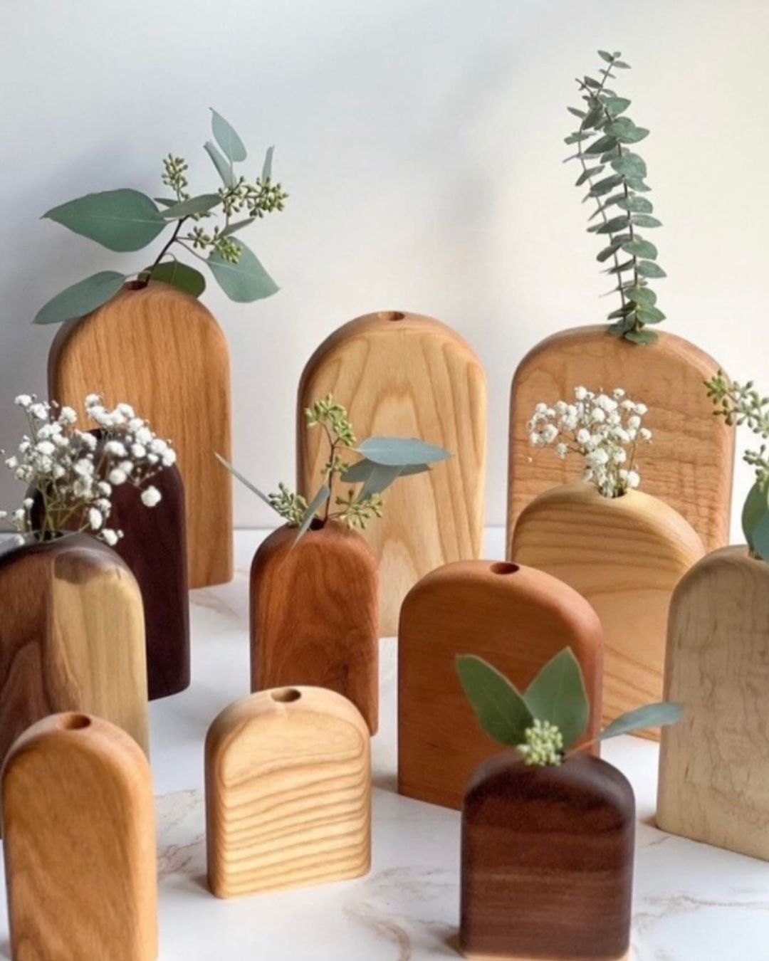 Collection of wooden bud vases of various sizes and wood types, each holding a sprig of greenery or flowers.