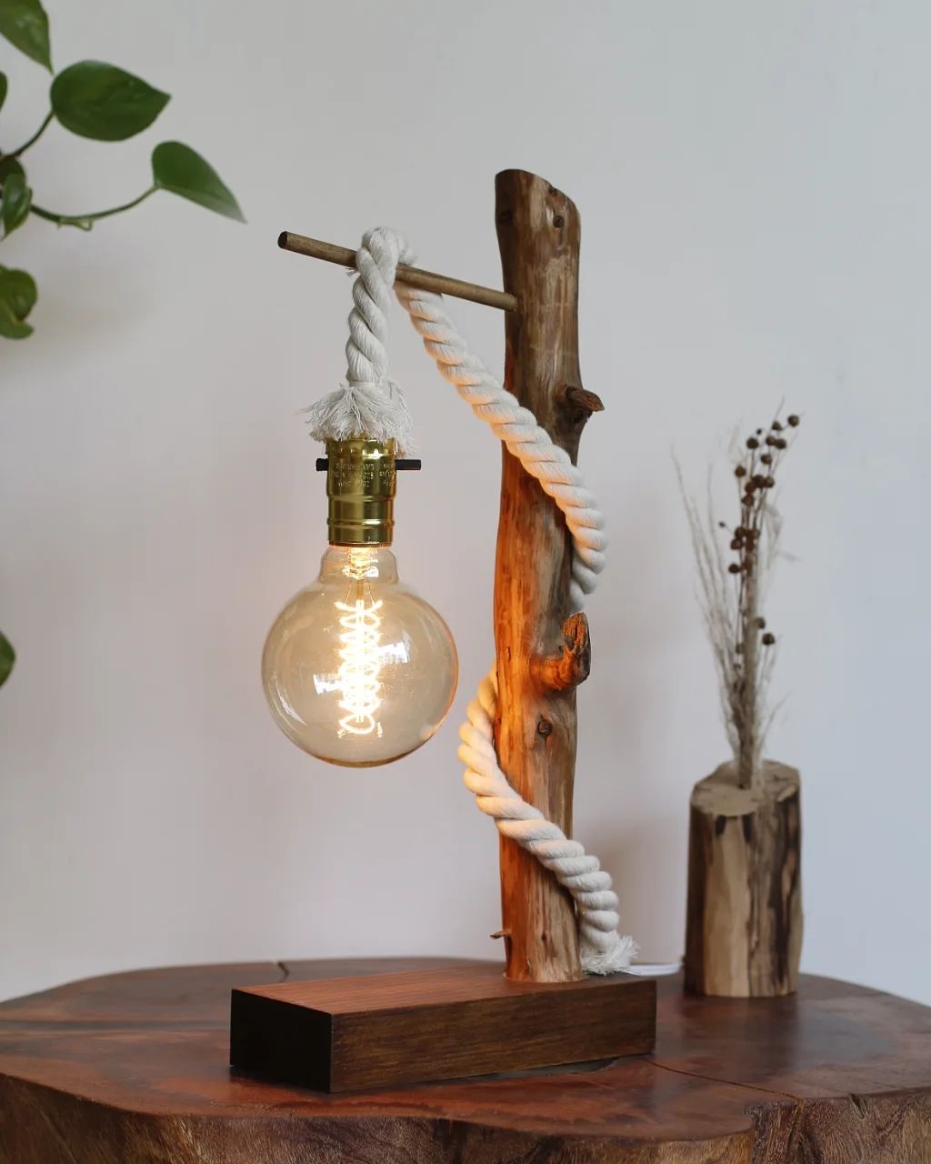 A unique wooden table lamp featuring a rustic branch, a thick rope gracefully looping over a metal bar, and an Edison bulb emitting a warm glow.