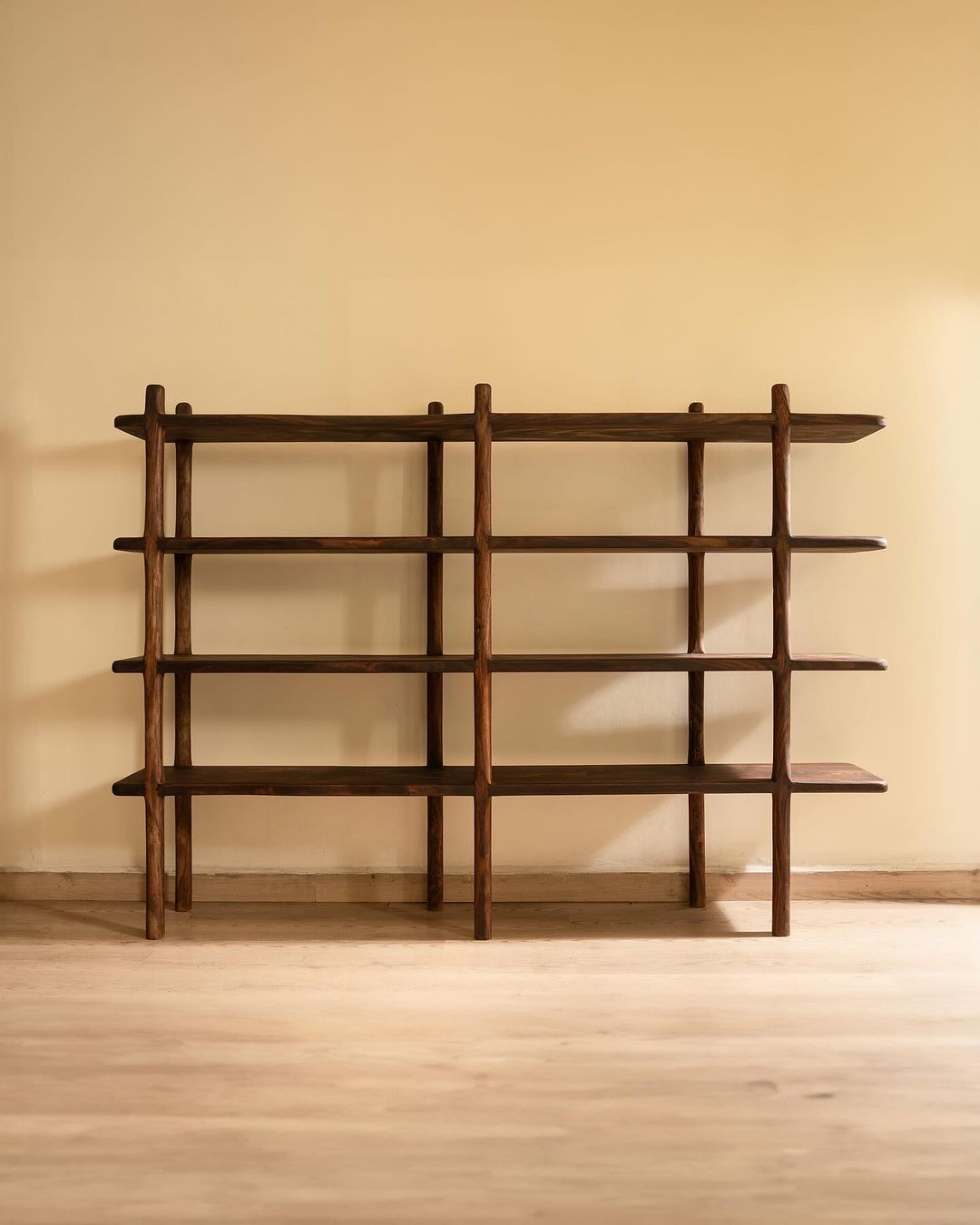 A minimalist wooden bookshelf structure with symmetric shelving against a warm beige wall.