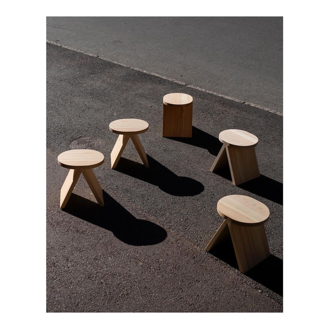 A modern and simplistic outdoor seating arrangement featuring four wooden three-legged stools on an asphalt surface