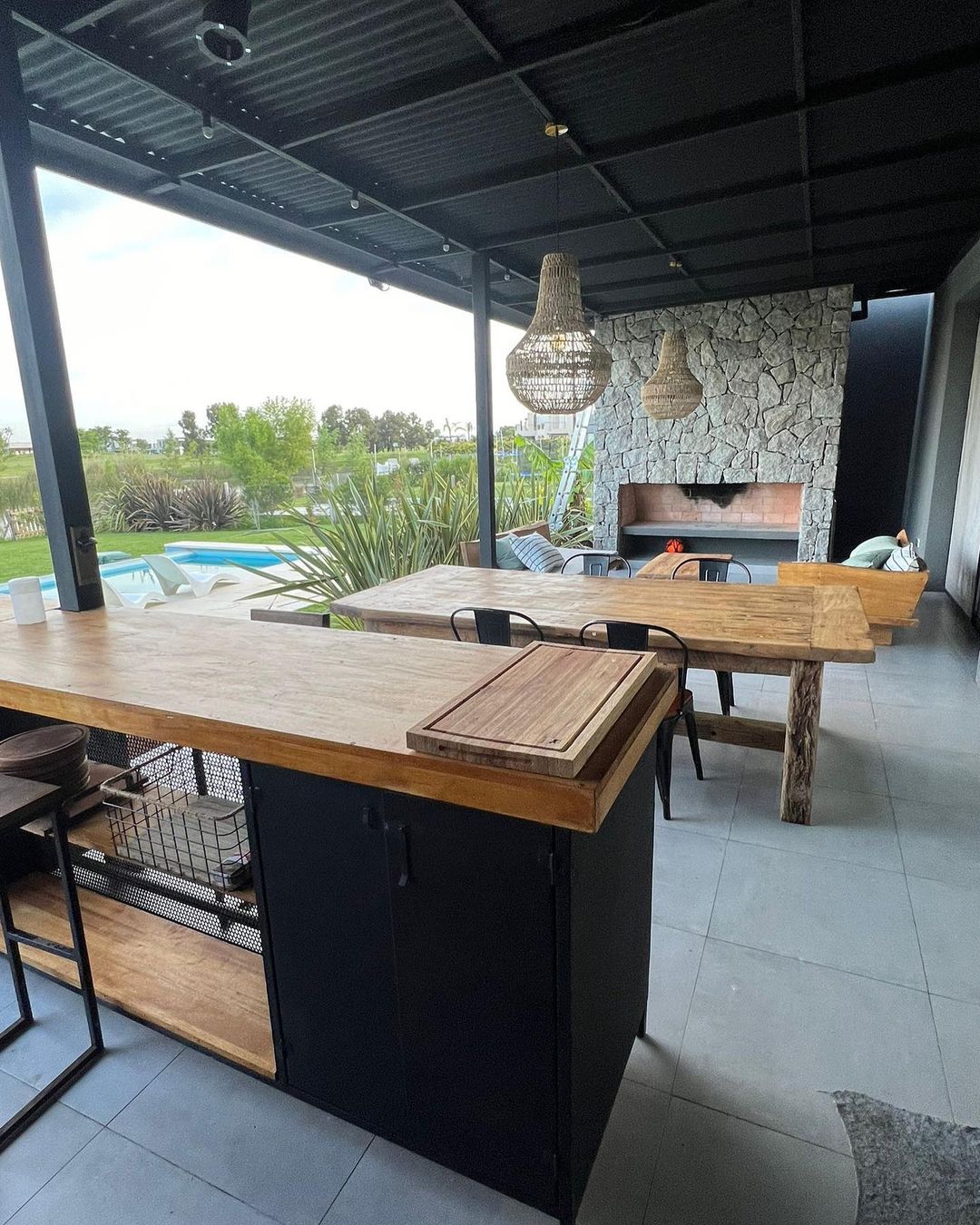 An inviting open-air patio area with a stone fireplace and wooden furnishings overlooking a pool