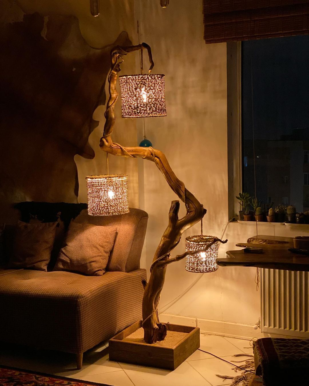 A rustic and artistic floor lamp made of natural twisted wood branches, holding two woven lampshades that cast a warm, patterned glow.