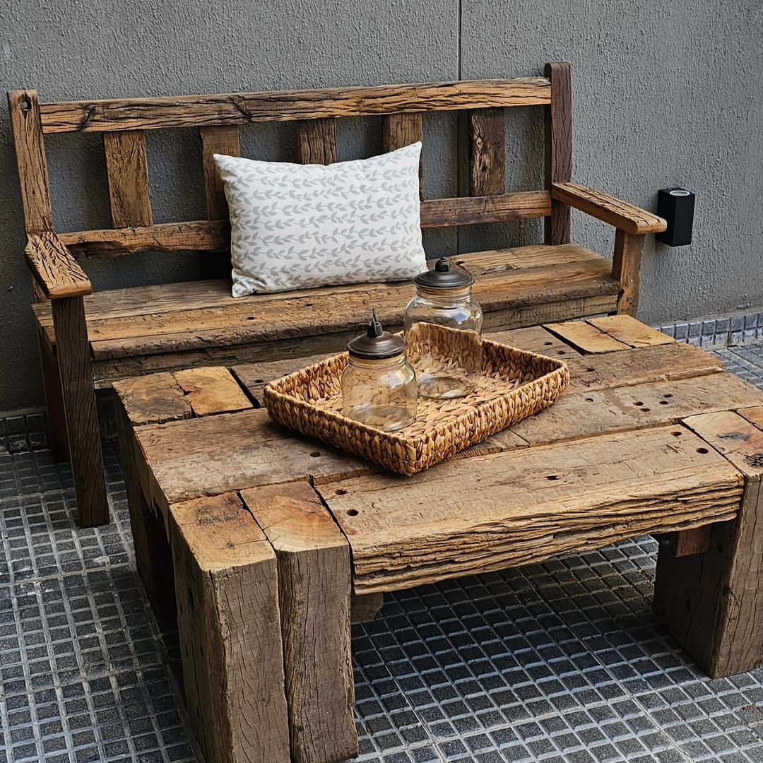 A rustic outdoor sitting area featuring a rugged wooden bench with a patterned throw pillow and a low reclaimed wood table adorned with a wicker tray and glass sugar and creamer set.