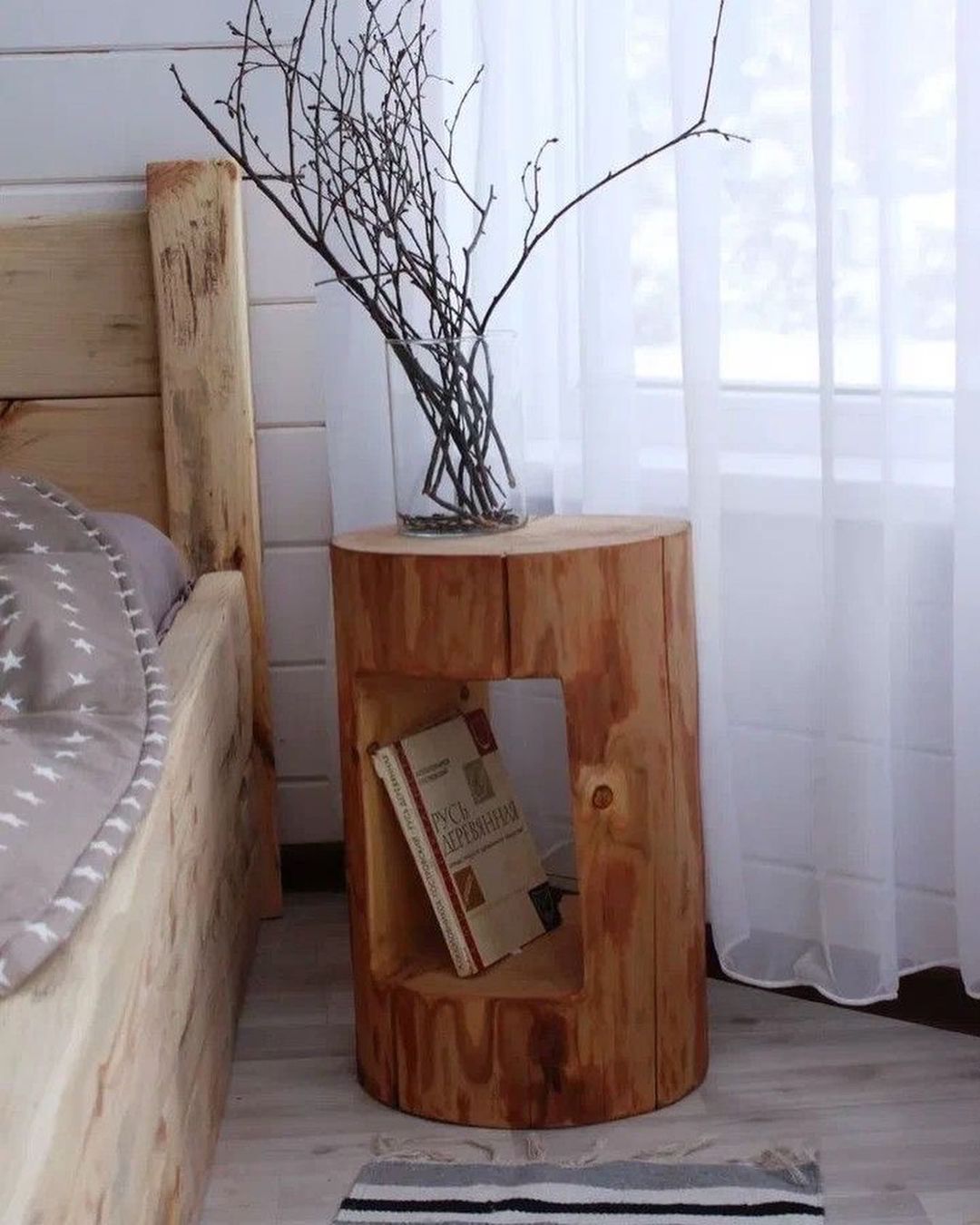 A rustic wood side table with an open shelf holding books and a glass vase with branches on top