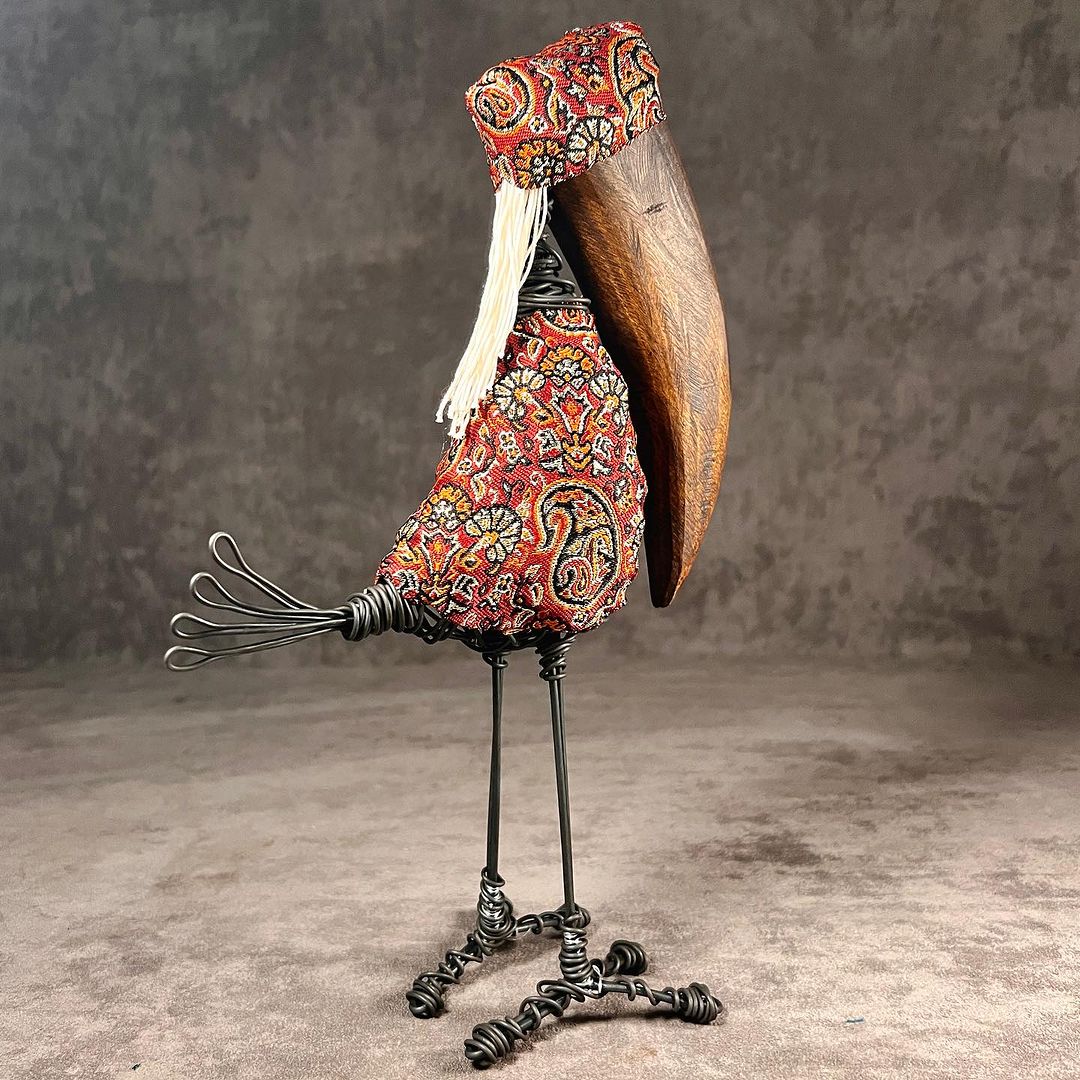 A whimsically crafted bird figure with wire legs, a patterned fabric body, and a wooden wing