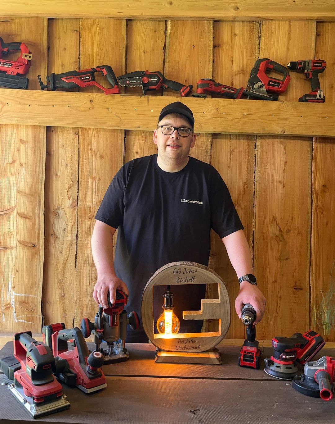 A man proudly posing with his woodworking tools arranged on a wooden wall and a handcrafted lamp with a lit Edison bulb